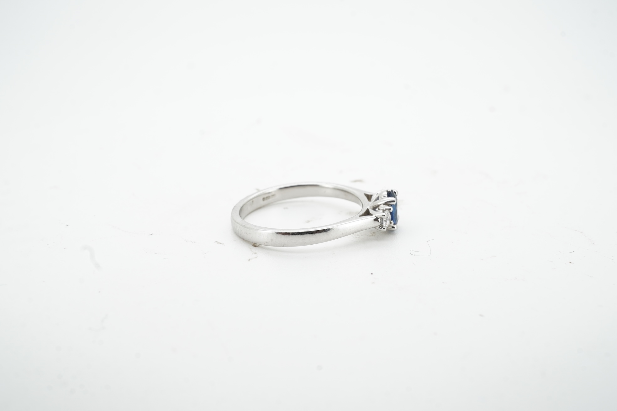 A sapphire and diamond three-stone ring, claw-set with an oval sapphire weighing approximately 0.35 carats, between shoulders set with brilliant-cut diamonds totalling approximately 0.12 carats, mounted in 9ct white gold